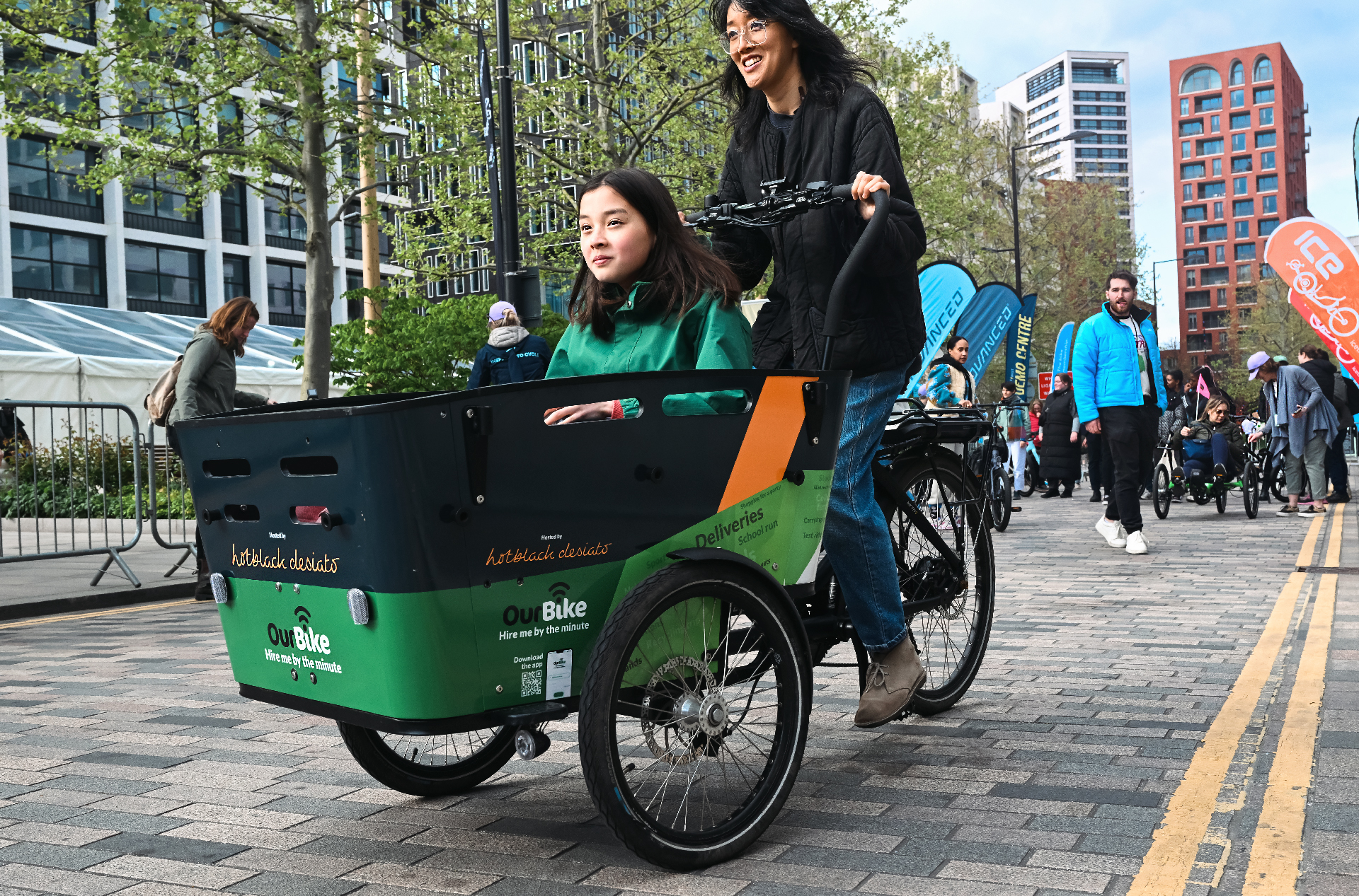 The image shows a mother and daughter enjoying test riding an e-cargo cycle at the Loud Mobility test ride track in central London. behind them are the flags of several event partners including Advanced, Brompton, Lime etc.